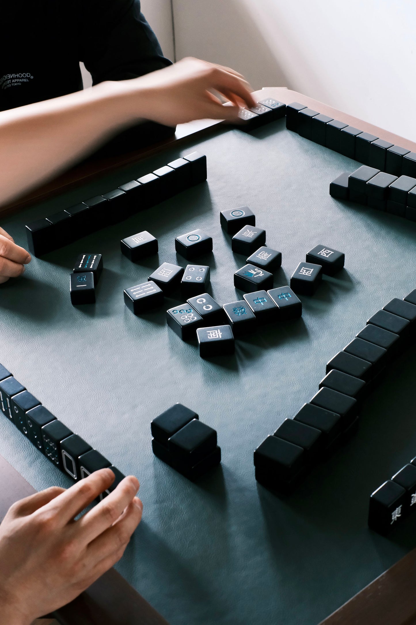 MJ88 Foldable Mahjong Dining Table – hands in action playing with black Mahjong tiles on green leather surface, showing real game use 麻雀餐桌,日本製,玩家雙手實際操作黑色麻雀牌,展現遊戲進行中的真實情境,綠色桌面襯托。