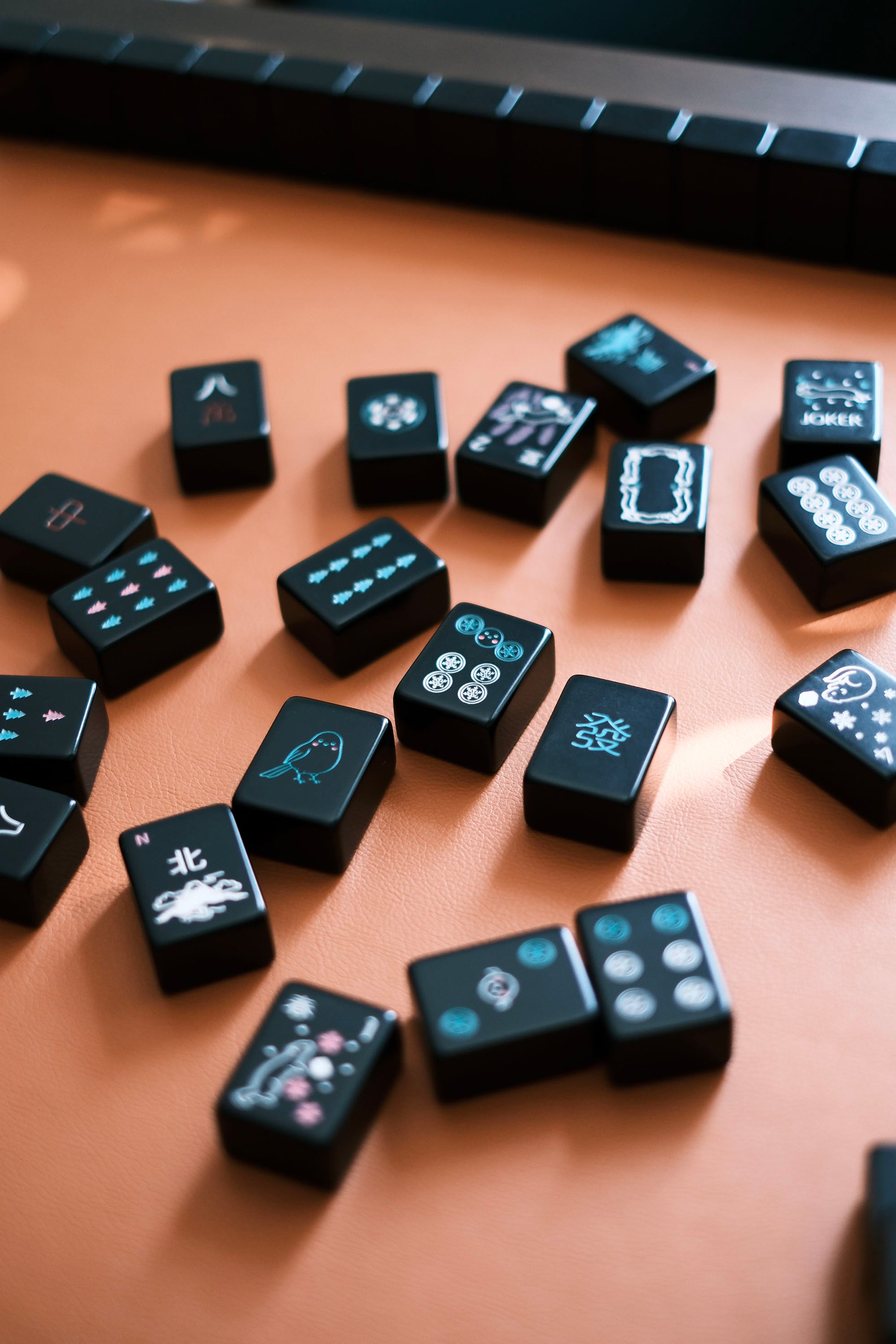 Black mahjong tiles with teal and pink Hokkaido seasonal motifs scattered on the table surface
黑色麻雀牌散落桌面，牌面為湖水綠與粉色北海道四季圖案
